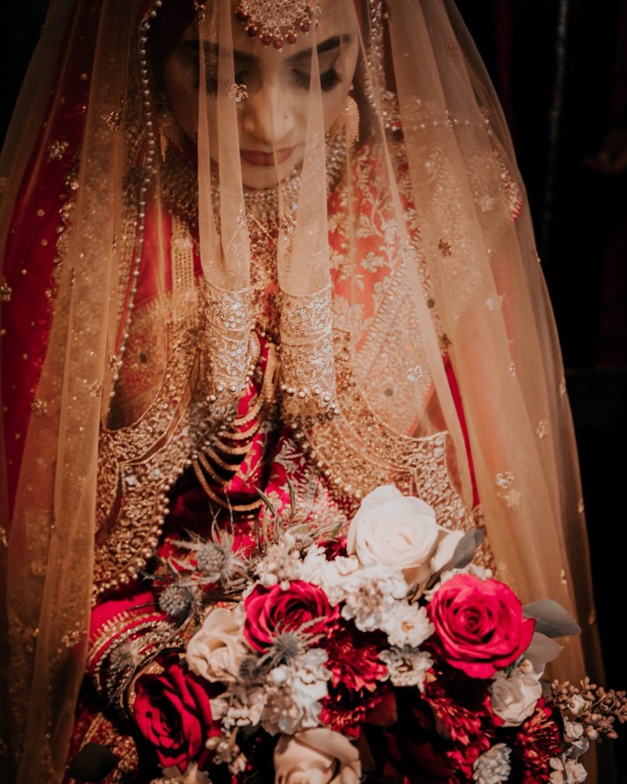 Veiled bride in red and gold attire holding a bouquet of red roses, white blooms, and blue thistle