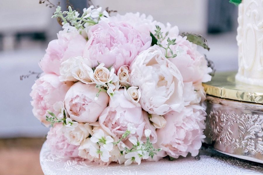 Blush peony and cream spray rose bridal bouquet resting beside a silver cake stand and wedding cake