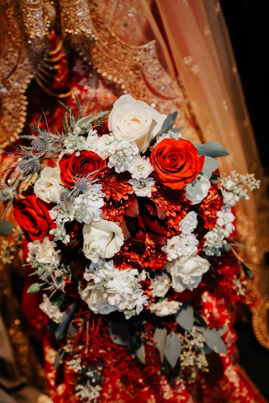 Bride in red and gold bridal attire holding a bouquet of red and white roses with blue thistle