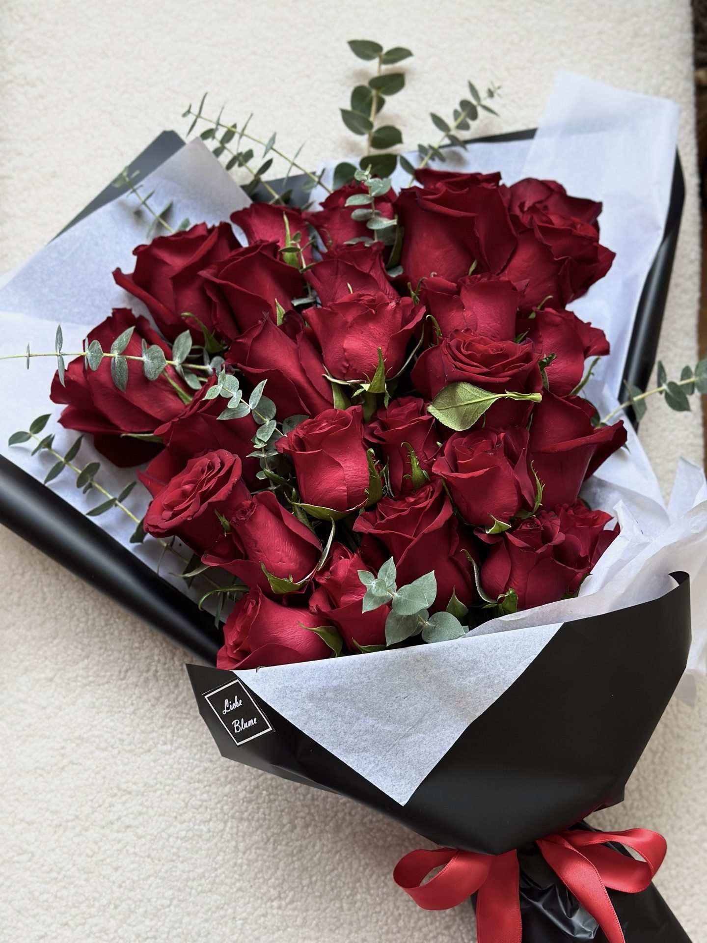 Red rose bouquet with blue eucalyptus in black Liebe Blüme wrap and gold-logo tag on cream boucle
